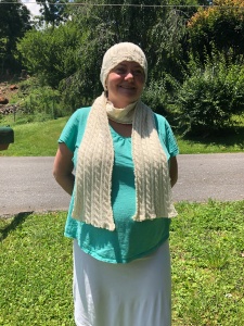 Photo of me wearing the Palindrome Hat and Scarf with green trees in the background - The scarf and hat are off-white with cables and ribbing.